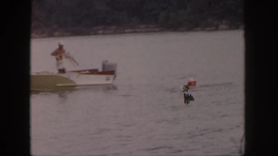 Faded 8mm frame from Michigan in 1961 showing a small boat, a standing man, and a red buoy on a quiet lake.
