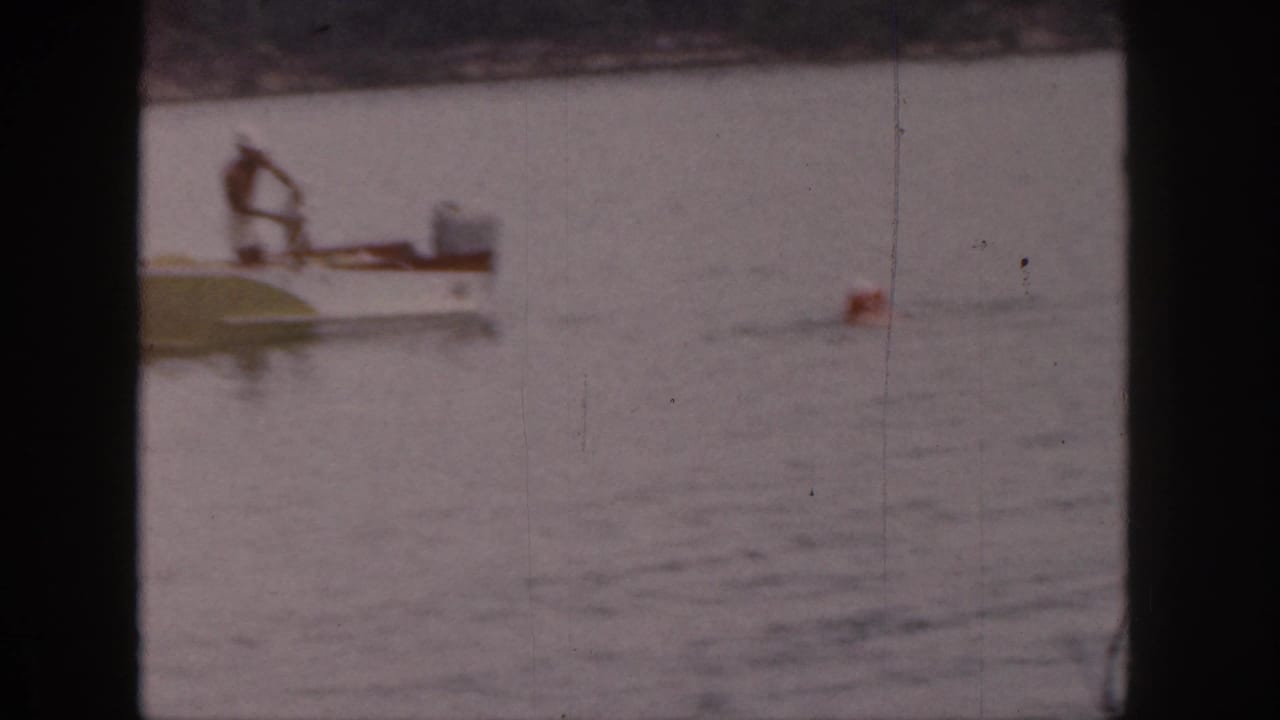 Man bends over a small motorboat on calm water with a red buoy nearby.