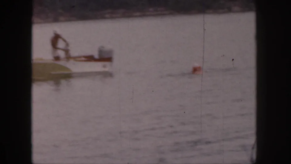 In 1961 Michigan home-movie footage, a man leans over a small motorboat on a calm lake near a red buoy.