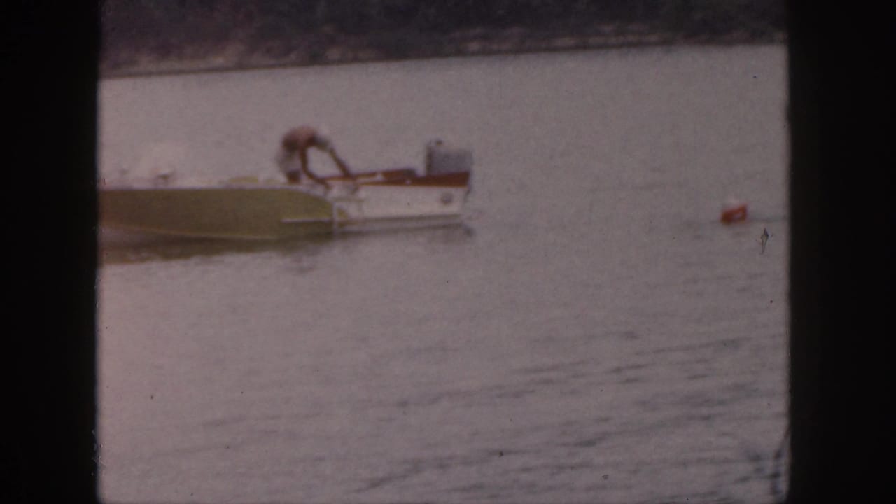 Small motorboat on a lake as a man leans down near the side and a red buoy floats nearby.