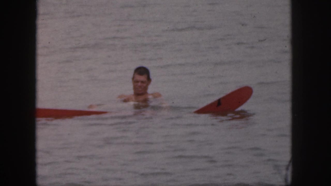 In 1961 Michigan home-movie footage, a shirtless man floats in calm water near a partially submerged red board.