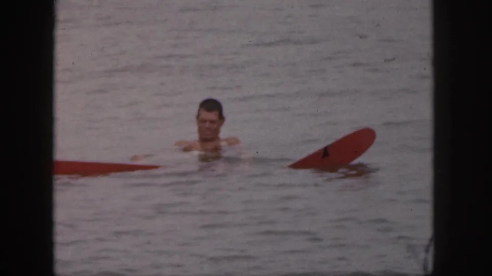 In 1961 Michigan home-movie footage, a shirtless man floats in calm water near a partially submerged red board.