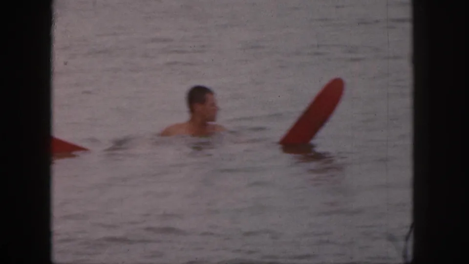 The man turns in the water near an upright red surfboard or float board in the 1961 Michigan home-movie scene.