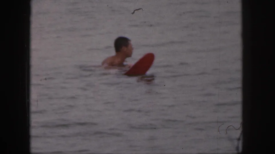 A shirtless man appears in profile in calm Michigan water, close to a red board, in faded 8mm color.