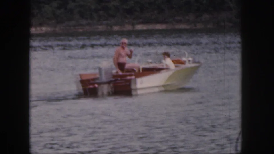1961 Michigan 8mm frame of a small wooden motorboat crossing lake water with two visible passengers and a distant shoreline.
