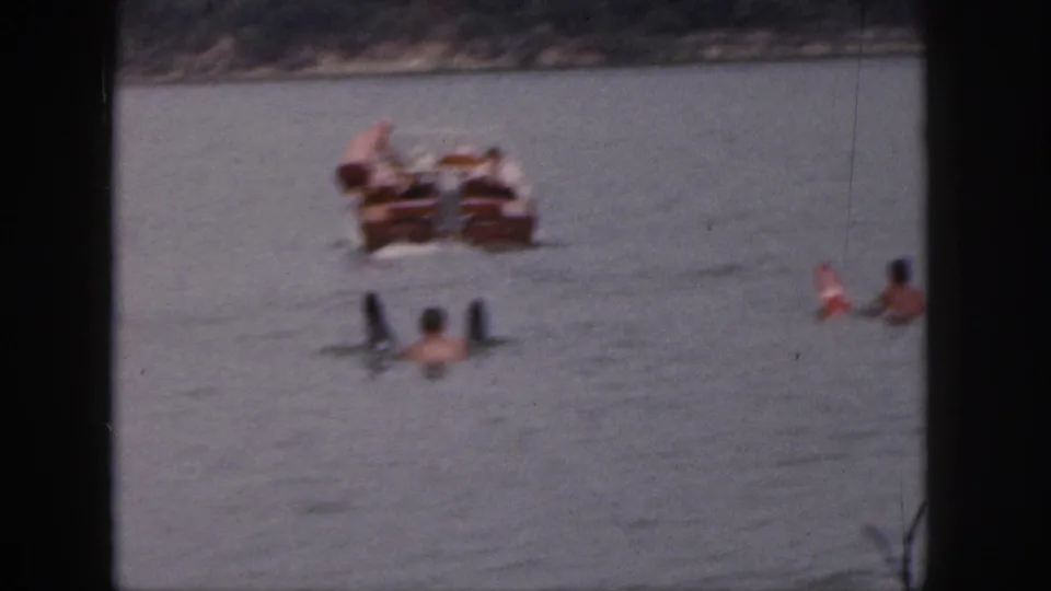 1961 Michigan home-movie frame showing swimmers spread across the lake near a wooden motorboat.