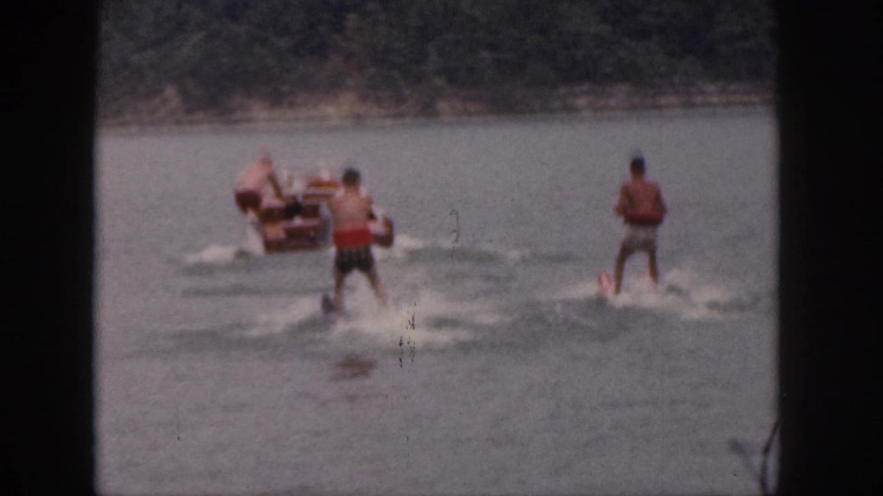 1961 Michigan home-movie frame showing two tandem water skiers behind a small motorboat on a wooded lake.