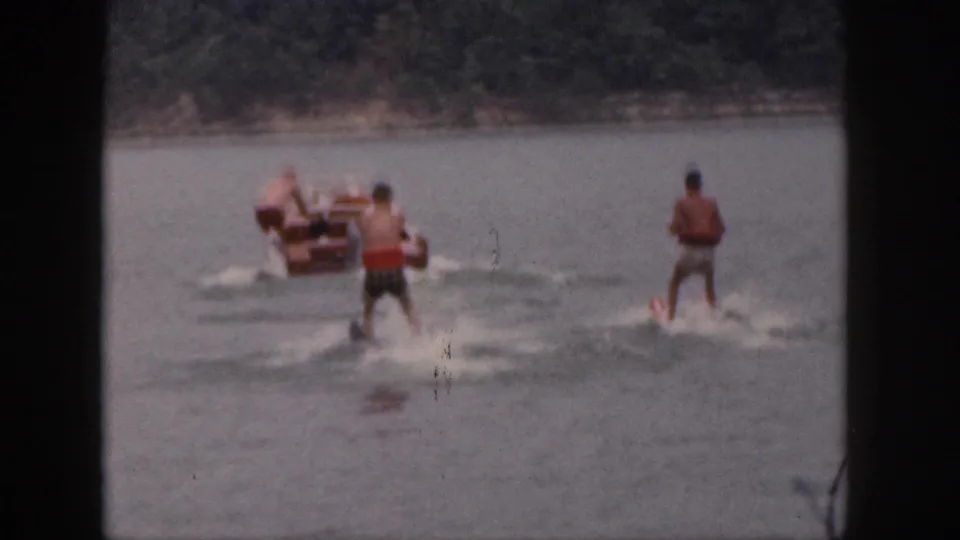 1961 Michigan home-movie frame showing two tandem water skiers behind a small motorboat on a wooded lake.