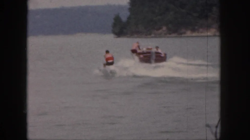 1961 Michigan home-movie frame showing skiers at different distances behind a motorboat near a wooded point.