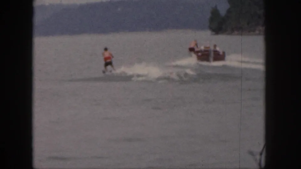 1961 Michigan 8mm frame with a skier crossing the wake behind a motorboat on a broad wooded lake.