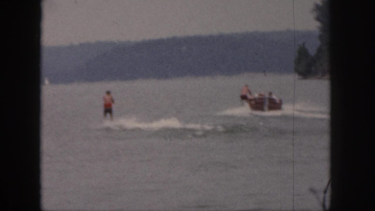 1961 Michigan 8mm frame showing a waterskier in a red top behind a small motorboat on a lake, with distant trees and soft film edges.