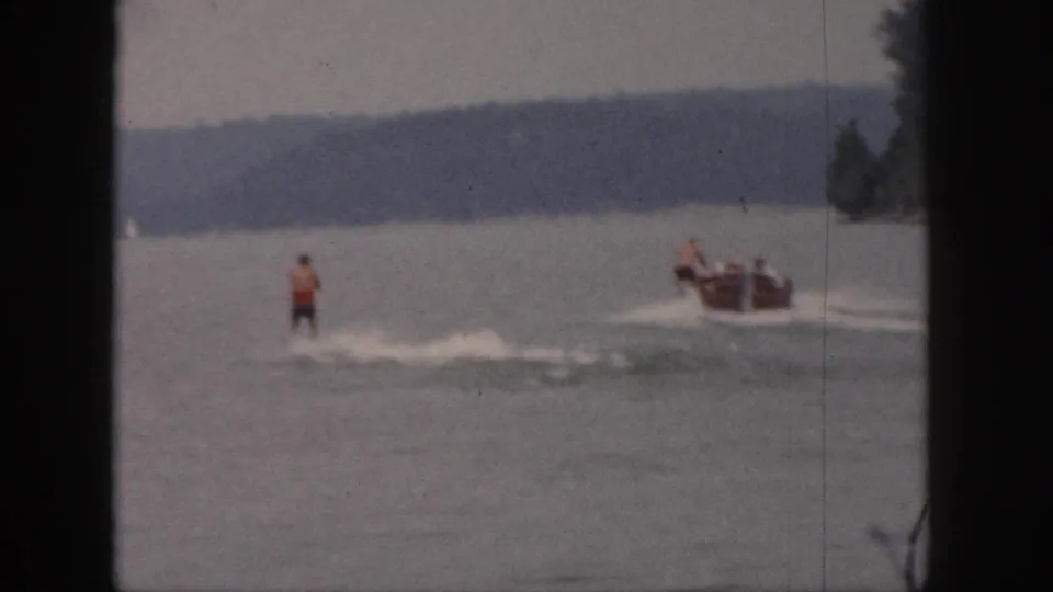 1961 Michigan 8mm frame showing a waterskier in a red top behind a small motorboat on a lake, with distant trees and soft film edges.