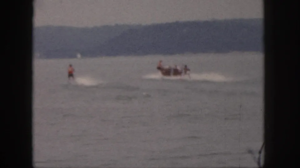 1961 Michigan 8mm frame with a waterskier and small passenger boat moving across a lake under soft, grainy exposure.