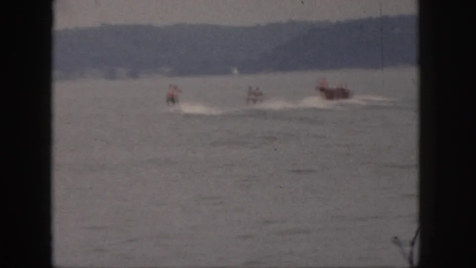 1961 Michigan home-movie frame showing a motorboat pulling waterskiers across a lake, seen from shore with dark 8mm borders.