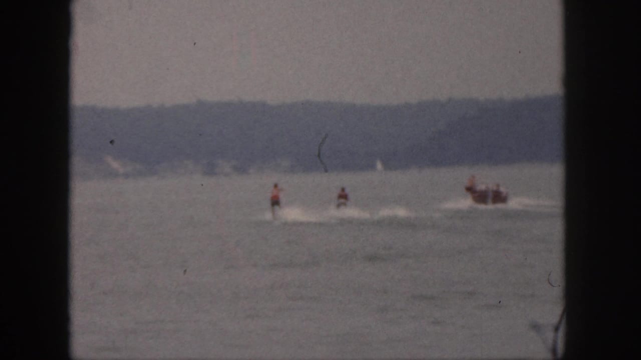 1961 Michigan home-movie frame showing distant riders on motorized watercraft with a following boat and white wakes on a hazy lake.