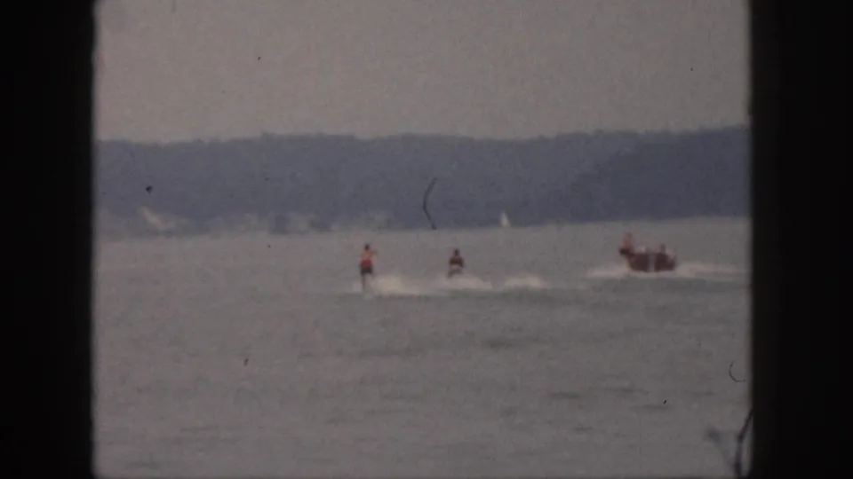 1961 Michigan home-movie frame showing distant riders on motorized watercraft with a following boat and white wakes on a hazy lake.
