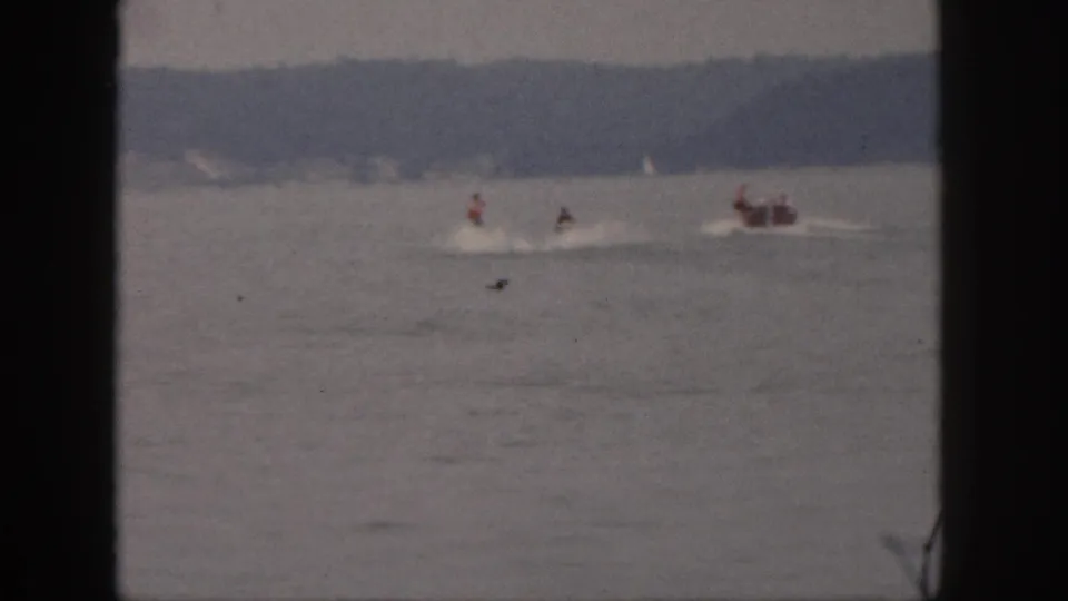Distant riders and a small motorboat create pale wakes on a Michigan lake in soft 1961 8mm home-movie footage.