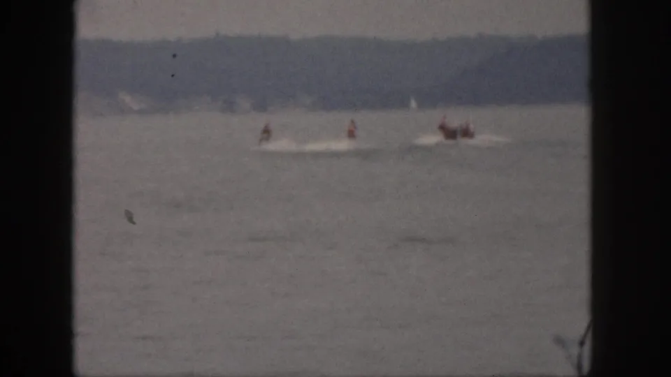 Soft-focus 8mm frame from Michigan in 1961 showing riders and a small boat moving left to right across calm lake water.