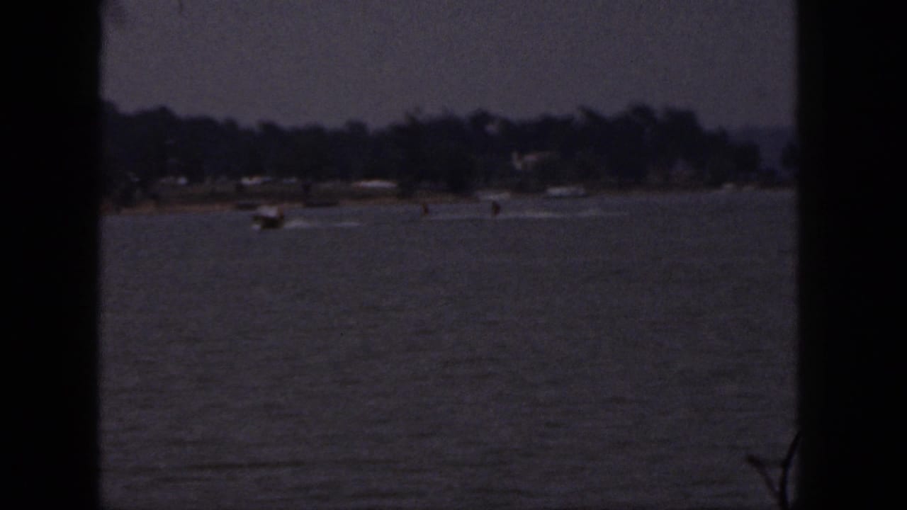 1961 Michigan 8mm frame showing a distant speedboat pulling water skiers across a lake near a tree-lined shore.