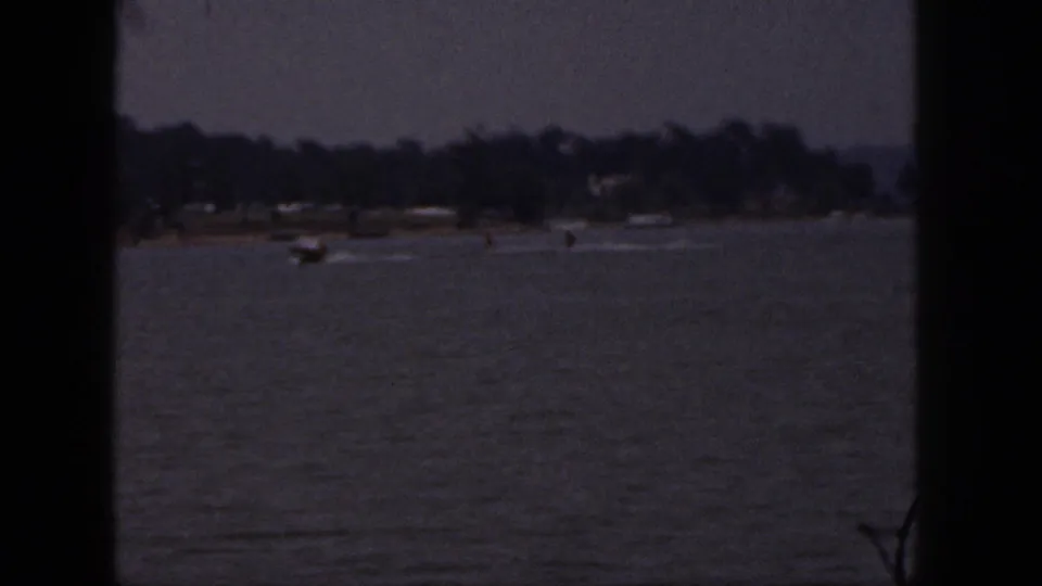 1961 Michigan 8mm frame showing a distant speedboat pulling water skiers across a lake near a tree-lined shore.