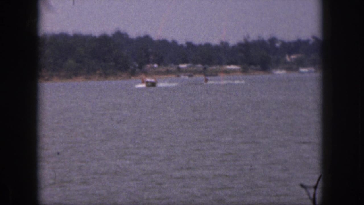 Speedboat pulls two water skiers near a wooded shoreline on a calm lake.