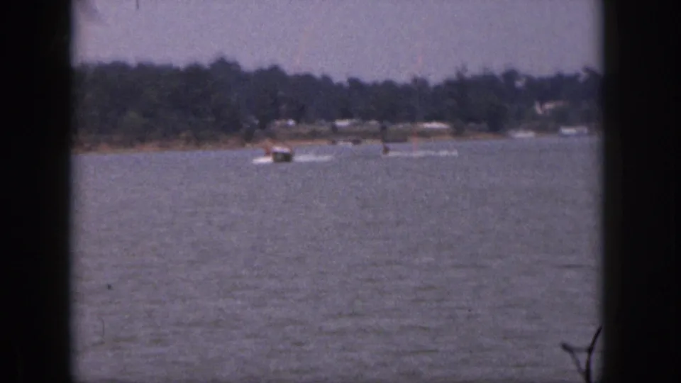 1961 Michigan home-movie frame with two distant water skiers following a speedboat across a calm lake.