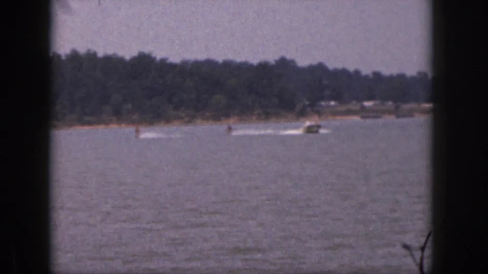 1961 Michigan home-movie frame with a speedboat and two skiers cutting white wakes across the lake.