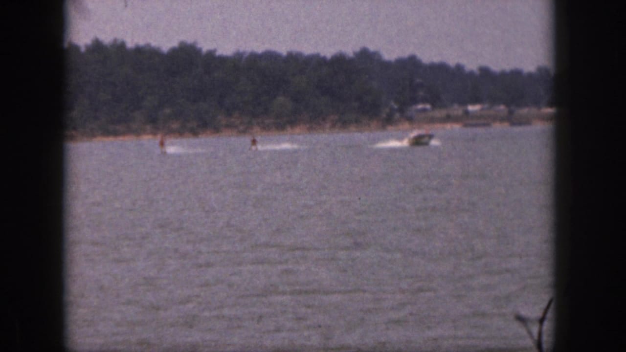 1961 Michigan 8mm frame showing distant water skiers and motorboats crossing a lake with a wooded shore beyond.