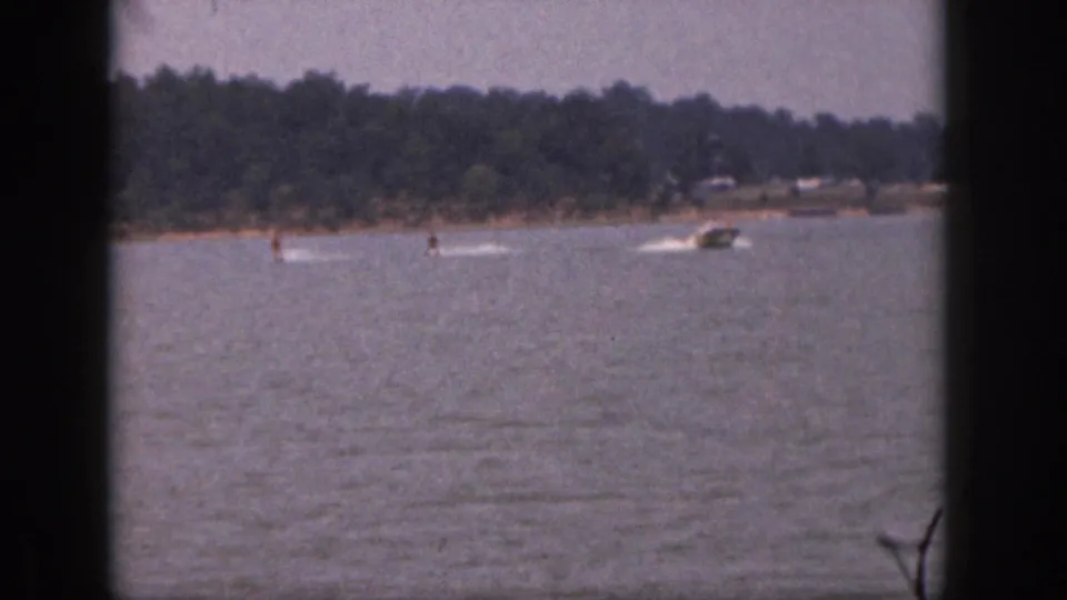 1961 Michigan 8mm frame showing distant water skiers and motorboats crossing a lake with a wooded shore beyond.