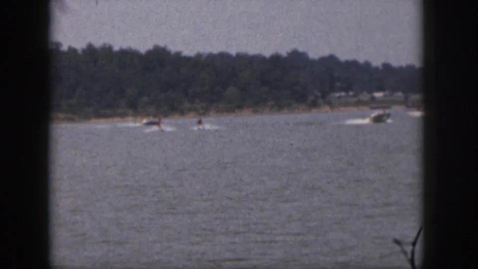 Distant water skiers and two small boats move across a Michigan lake in 1961 home-movie footage.