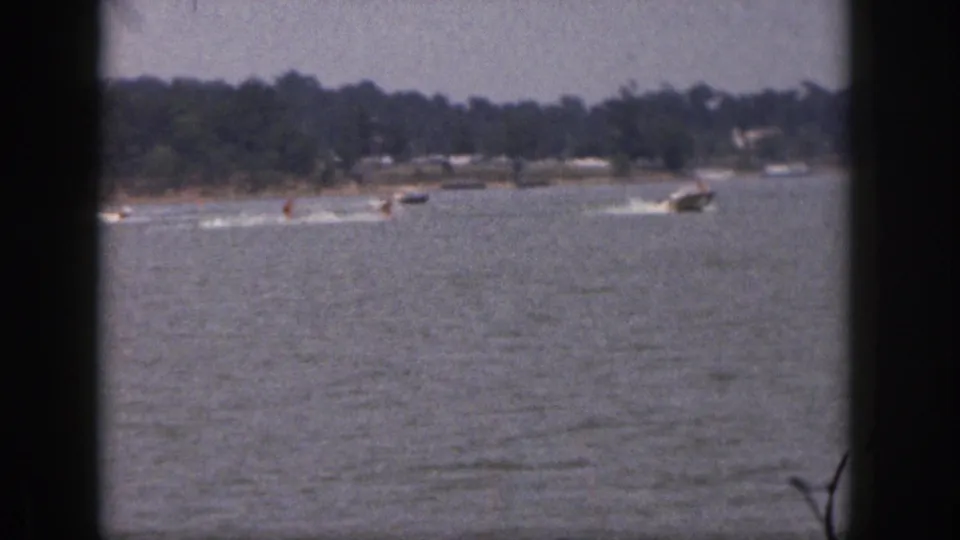Faded 8mm frame of Michigan lake recreation in 1961, with skiers, boats, wakes, and a wooded shore.