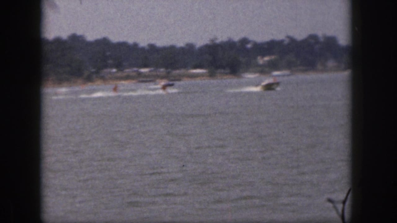 1961 Michigan 8mm frame showing a distant motorboat towing two skiers across a lake with shoreline trees beyond.