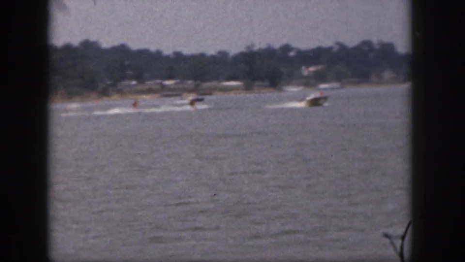 1961 Michigan 8mm frame showing a distant motorboat towing two skiers across a lake with shoreline trees beyond.
