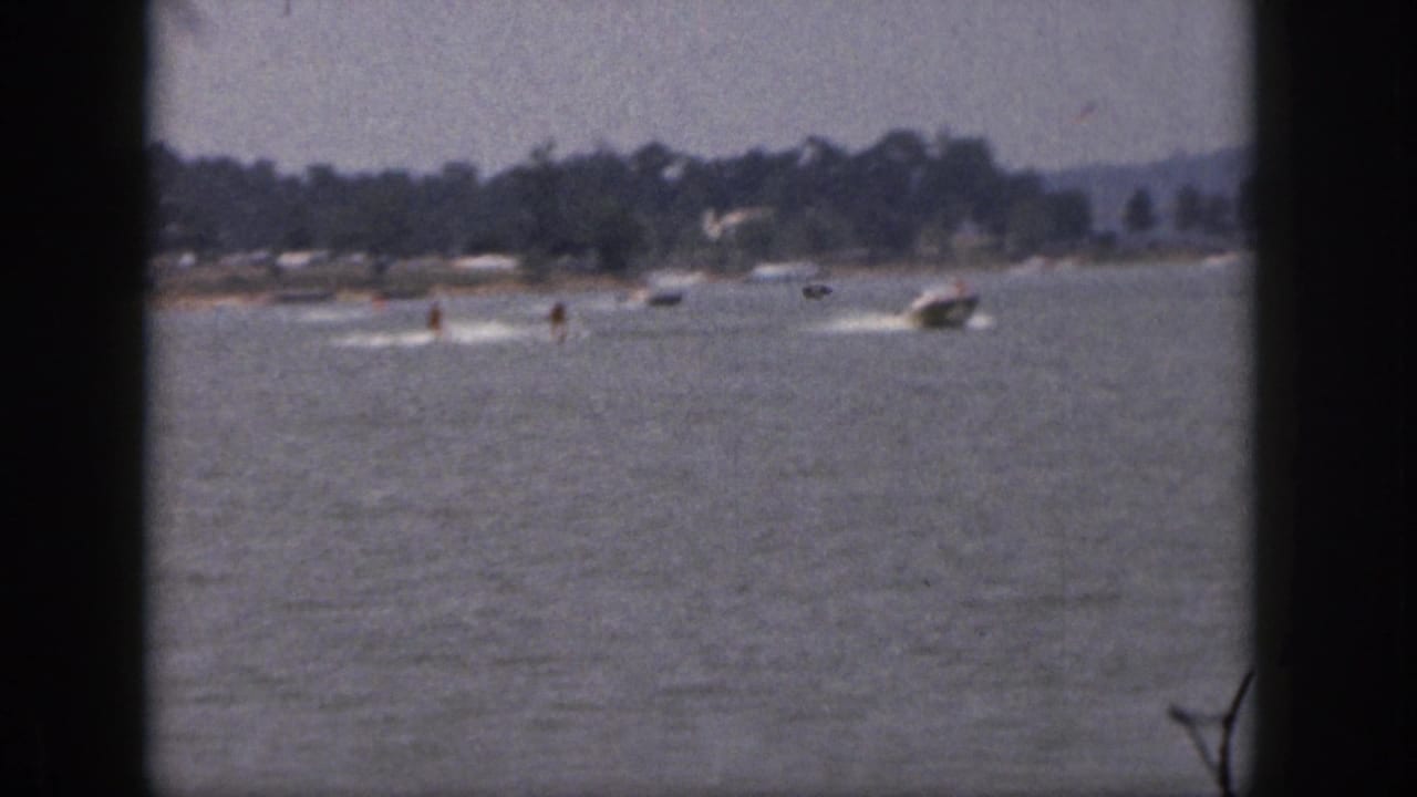 Distant 1961 Michigan lake view with a motorboat towing skiers, framed by dark 8mm borders and a tree-lined shore.