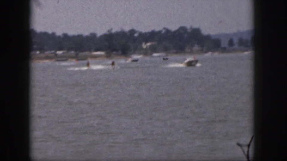 Distant 1961 Michigan lake view with a motorboat towing skiers, framed by dark 8mm borders and a tree-lined shore.