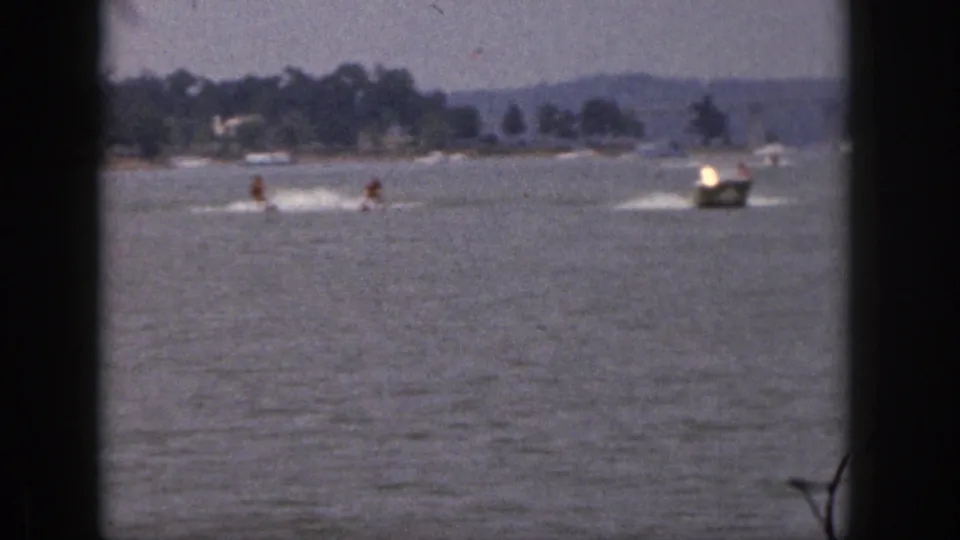 Soft-focus 8mm frame from Michigan in 1961 showing a motorboat pulling two skiers over a lake with wooded hills behind.