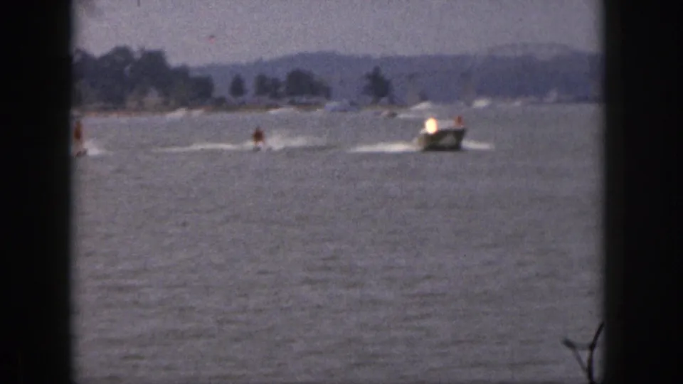 1961 Michigan home-movie frame with a motorboat towing two waterskiers across open water near a wooded shoreline.