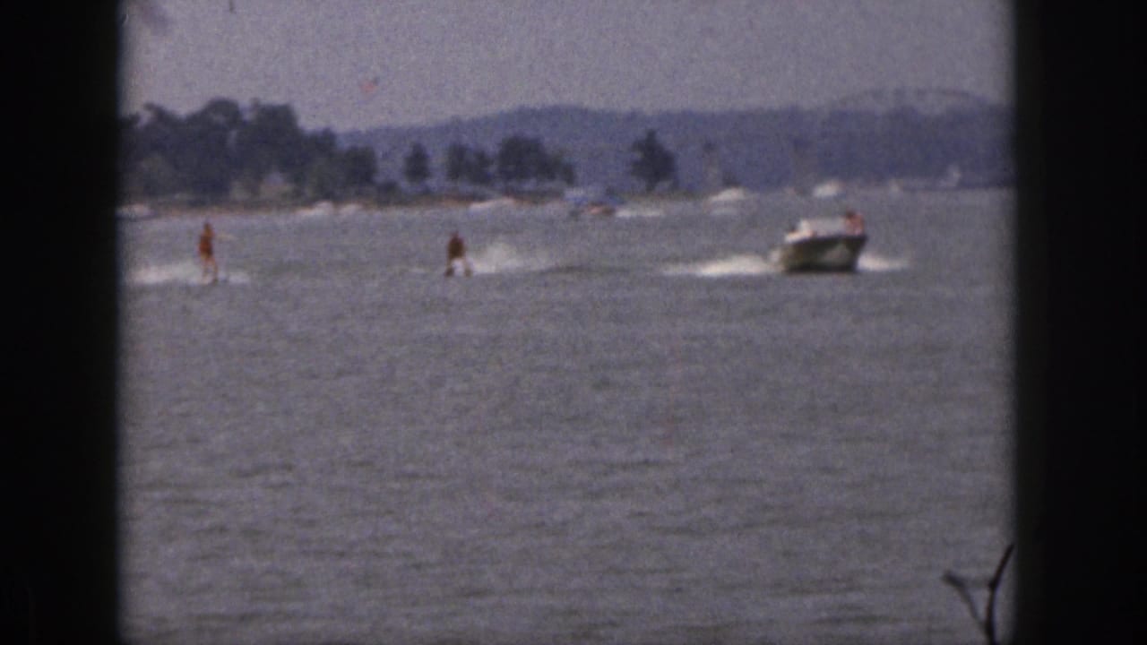 1961 Michigan 8mm frame showing two distant water skiers and a motorboat crossing open lake water near a tree-lined shoreline.