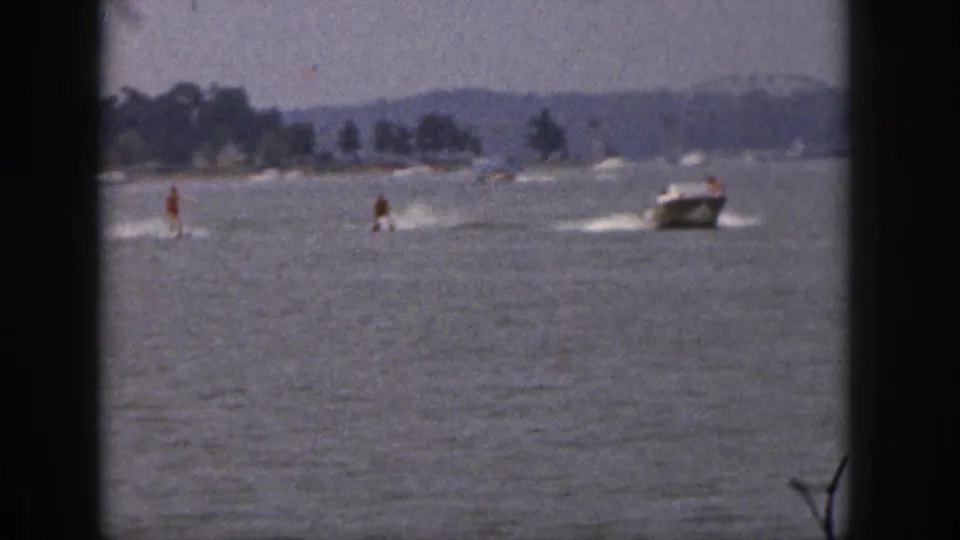 1961 Michigan 8mm frame showing two distant water skiers and a motorboat crossing open lake water near a tree-lined shoreline.