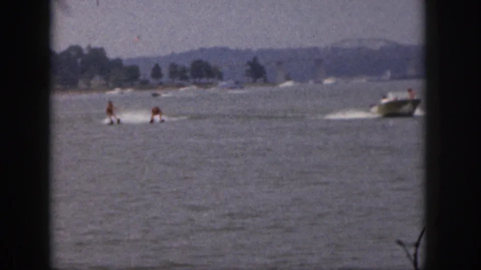 Distant 1961 Michigan lake view with two water skiers following a motorboat, framed by soft 8mm borders and a hazy shoreline.