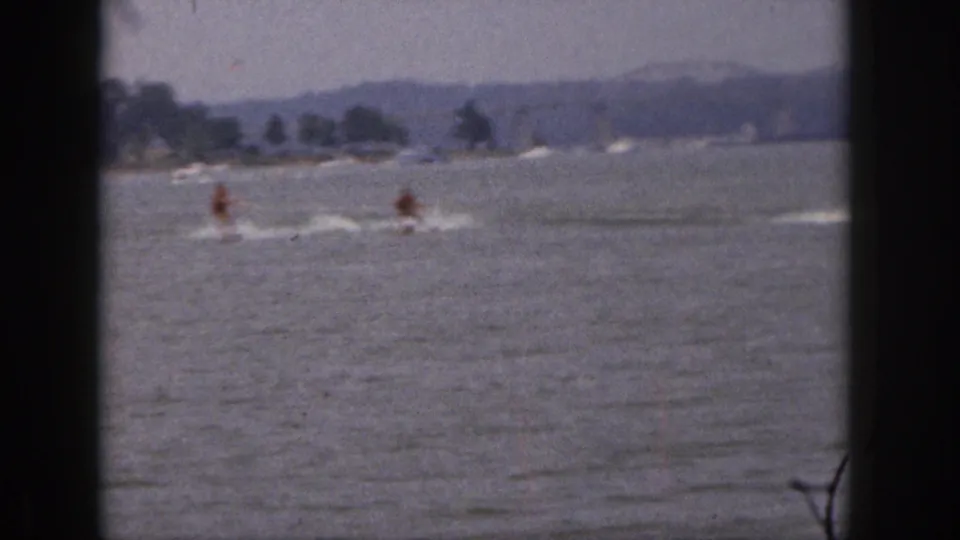 Soft 8mm home-movie frame from 1961 Michigan showing two distant skiers cutting across lake water with white spray behind them.
