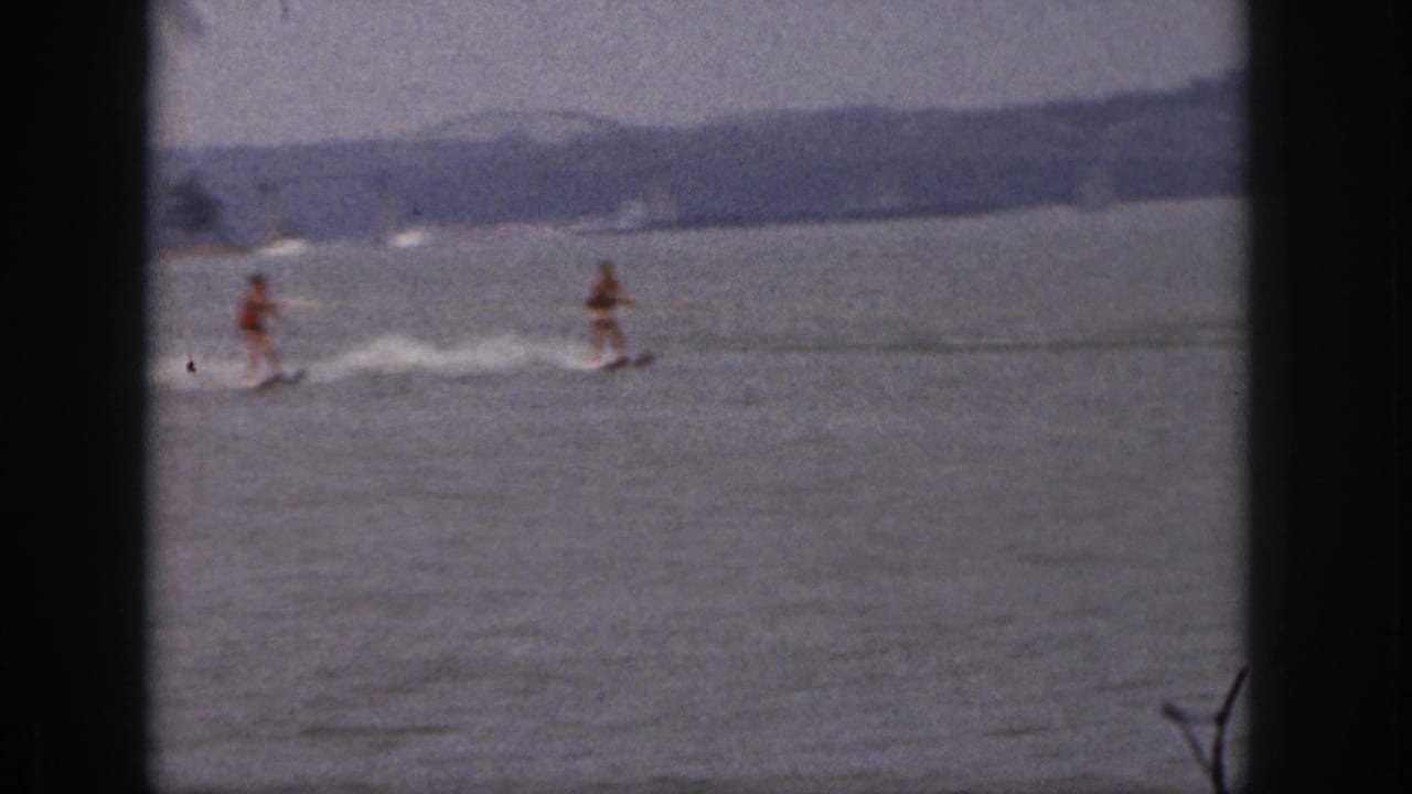 Two adult water skiers glide across a Michigan lake in 1961 home-movie footage, seen through dark 8mm frame borders.