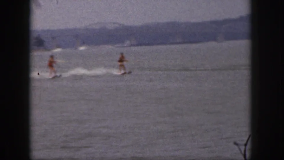 Two adult water skiers glide across a Michigan lake in 1961 home-movie footage, seen through dark 8mm frame borders.