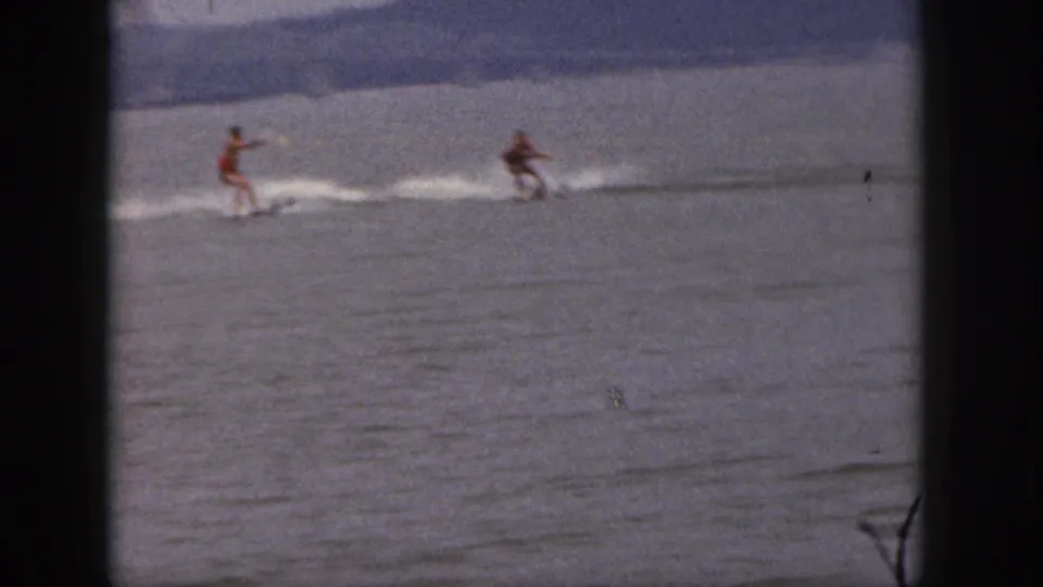 Two adult skiers are pulled across a calm Michigan lake, with soft grain and faded color from the 8mm source.