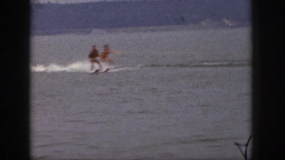 Two adult water skiers move close together across the lake in grainy 1961 Michigan home-movie footage.