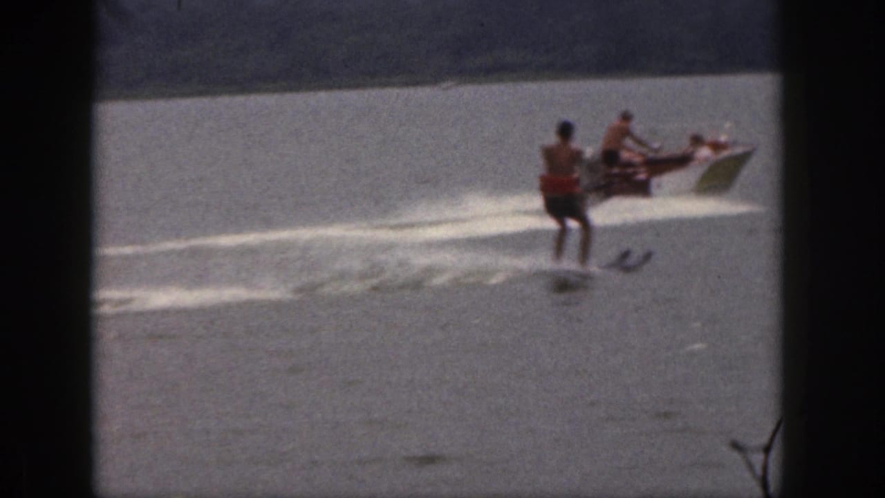 1961 Michigan home-movie frame showing a water skier trailing a small motorboat on a calm lake with a tree-lined shore behind.