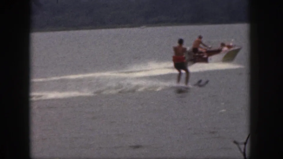 1961 Michigan home-movie frame showing a water skier trailing a small motorboat on a calm lake with a tree-lined shore behind.