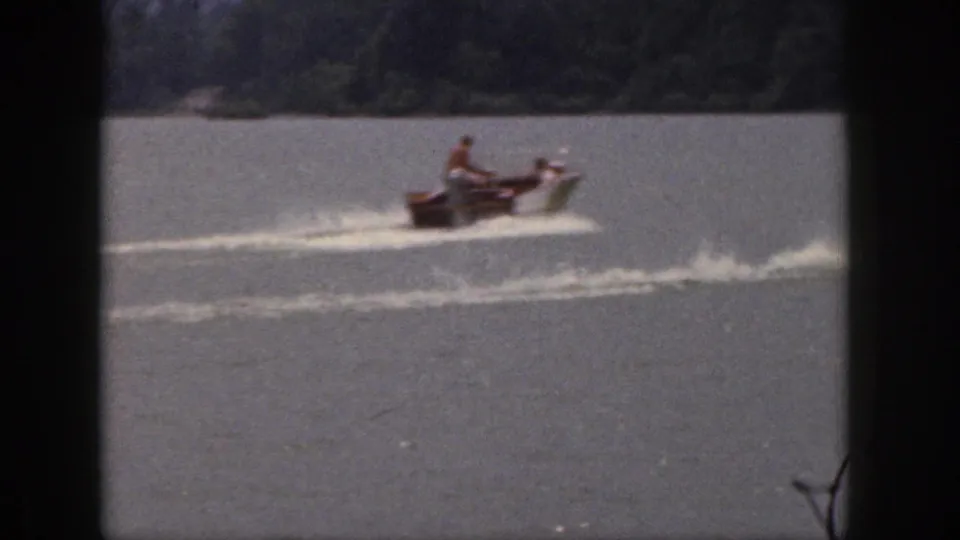 A small motorboat moves across a Michigan lake in 1961, with passengers visible and a long wake trailing across the water.