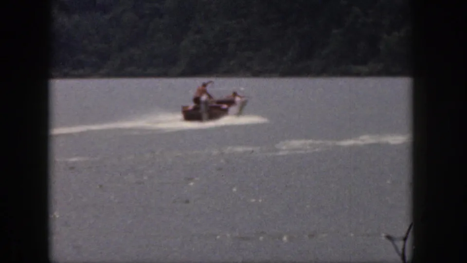 1961 Michigan lake scene with a small motorboat crossing left to right, wake spreading behind it and shoreline trees in the background.