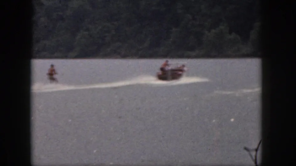 Distant 1961 Michigan home-movie view of a water skier following a small motorboat across a calm lake with a wooded shoreline.
